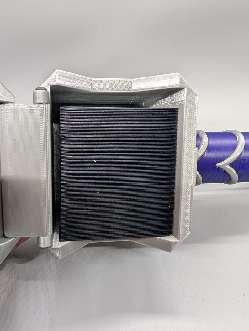 May include: A close-up of a 3D-printed object with a silver-gray frame and a black, ridged interior. The object has a purple and silver handle. The design appears to be a stylized weapon or tool.