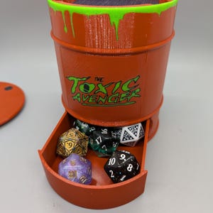 May include: Orange barrel-shaped dice holder with a green and black "The Toxic Avenger" logo. The top rim has green dripping paint. A drawer holds several multi-sided dice in various colors. A matching orange lid is visible.