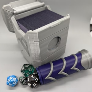 May include: A gray, 3D-printed card storage box with an open lid, revealing a stack of dark cards. A purple and silver dice holder is next to the box, with several multi-sided dice scattered nearby. The box has a circular opening.