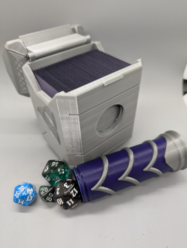 May include: A gray, 3D-printed card storage box with an open lid, revealing a stack of dark cards. A purple and silver dice holder is next to the box, with several multi-sided dice scattered nearby. The box has a circular opening.