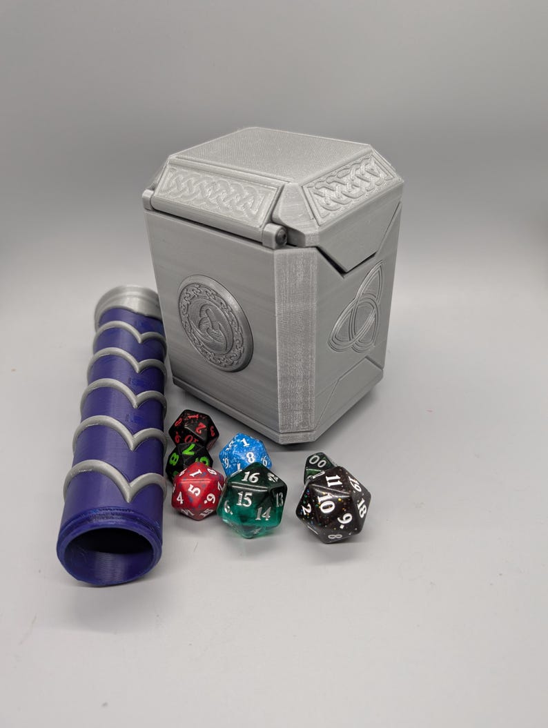 May include: A gray, 3D-printed dice box with Celtic knot and circular designs, a matching dice roller, and several multi-sided dice in various colors. The box is rectangular with a hinged lid.