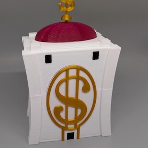 Könnte beinhalten: Eine weiße, 3D-gedruckte Geldkassette mit einer roten Kuppel und einem goldenen Dollarzeichen oben. Die Kassette hat ein goldenes Dollarzeichen auf der Vorderseite.