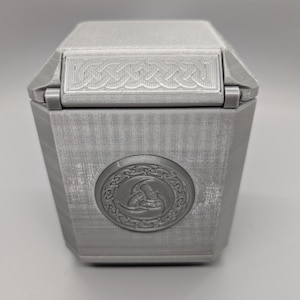 May include: A silver-colored, hexagonal box with a hinged lid. The lid features a decorative, woven-like design. A circular emblem with a detailed relief is centered on the front of the box. The box has a textured surface.