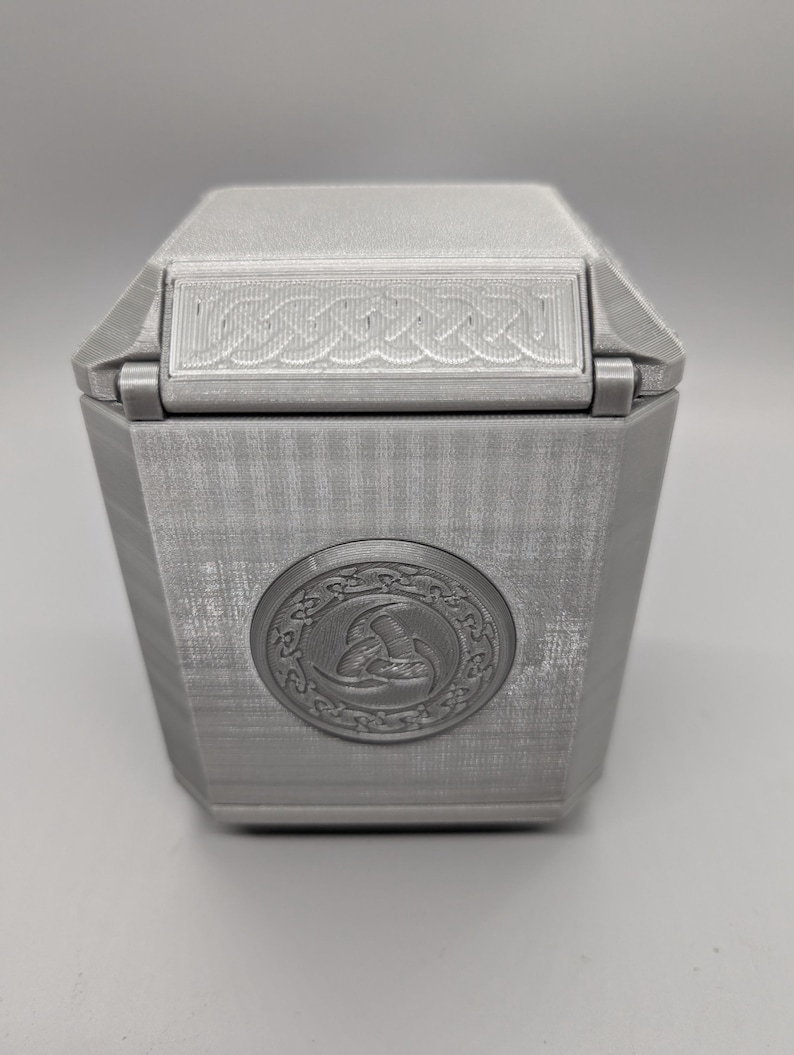 May include: A silver-colored, hexagonal box with a hinged lid. The lid features a decorative, woven-like design. A circular emblem with a detailed relief is centered on the front of the box. The box has a textured surface.
