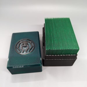 Puede incluir: Una caja de baraja de cartas verde y negra con un logotipo de araña en la parte delantera. La caja está abierta y las cartas son visibles en el interior.