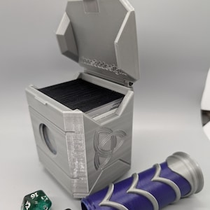 May include: A gray, 3D-printed card storage box with an open lid, a Celtic knot design, and a blue and silver dice roller. Several multi-sided dice are scattered nearby. The text "fane3ddesign" is visible on the lid.