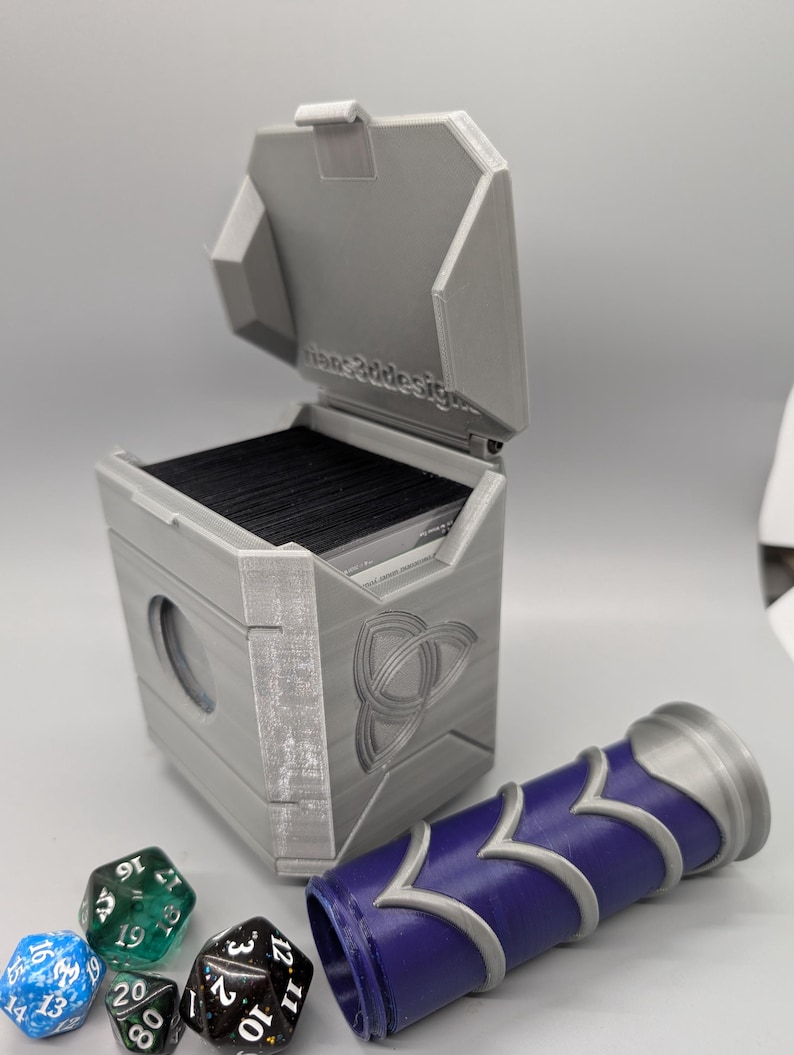 May include: A gray, 3D-printed card storage box with an open lid, a Celtic knot design, and a blue and silver dice roller. Several multi-sided dice are scattered nearby. The text "fane3ddesign" is visible on the lid.