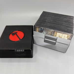 May include: A black and gray 3D printed box with a red circle and an axe symbol on the lid. The box is open and a stack of cards is visible inside. The cards are black and white with a gold border.