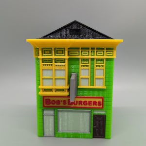 Puede incluir: Modelo impreso en 3D del restaurante Bob's Burgers. El edificio es verde con adornos amarillos, un tejado negro y un letrero rojo que dice "Bob's Burgers". El modelo tiene ventanas, una puerta y un letrero.
