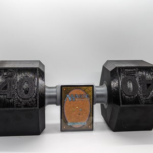 May include: A black dumbbell set with weights of 40 pounds on each side. The dumbbell is made of cast iron and has a hexagonal shape. A Magic: The Gathering card is in the center of the dumbbell.