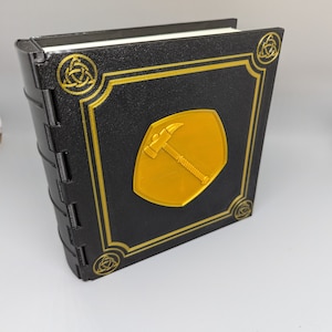 May include: A black book with a gold border and a gold emblem of a hammer and pickaxe on the front cover. The book has a Celtic knot design on the spine and corners.