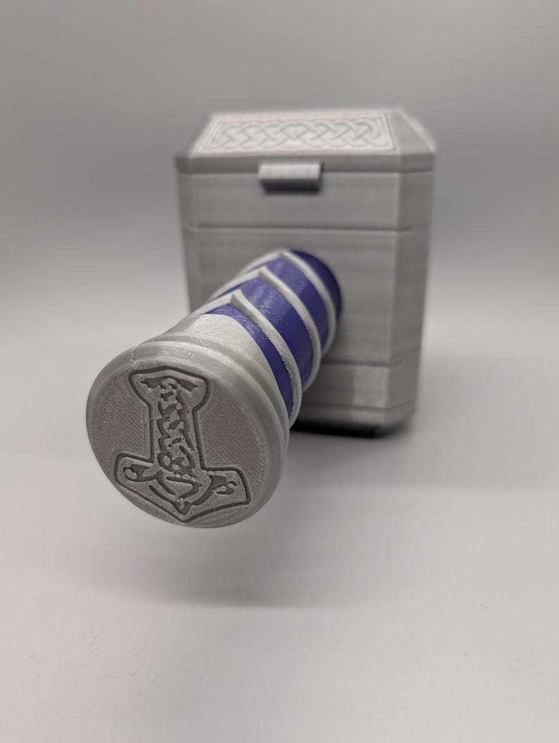 May include: A gray, 3D-printed replica of Thor's hammer, Mjolnir. The hammer features a cylindrical handle with blue and white stripes and a gray head with a detailed design. The head of the hammer has a circular end with a hammer symbol.
