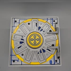 May include: A square mechanical vault door model featuring a circular silver and yellow rotating wheel with eight radial locking bars. The center displays a yellow circular emblem with four small blue gear icons. Blue gears are visible behind the silver frame, which is secured by black screws and a side hinge.