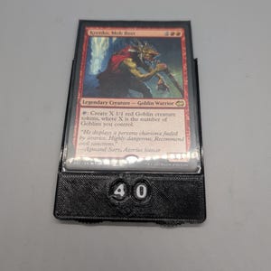 Peut inclure: Une carte Magic: The Gathering avec "Krenko, Mob Boss", un Guerrier Gobelin rouge. La carte est dans un support de carte noir avec le numéro "40" visible. L'illustration montre un gobelin avec une arme.