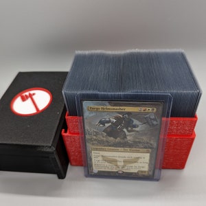 Puede incluir: Una caja de mazo roja y negra con una tapa transparente, que contiene una pila de cartas de juego. La carta superior es una carta de Magic: The Gathering con Zurgo Helmsmasher, una criatura legendaria con un esquema de color rojo, negro y dorado.