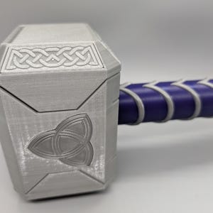 May include: A silver-colored hammer with a geometric design on the head and a purple handle. The hammer head features a Celtic knot design and a triquetra symbol. The handle is wrapped in white and purple material.