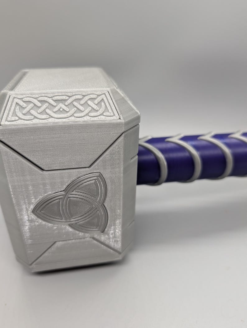 May include: A silver-colored hammer with a geometric design on the head and a purple handle. The hammer head features a Celtic knot design and a triquetra symbol. The handle is wrapped in white and purple material.