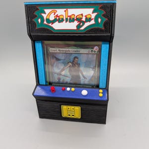 Puede incluir: Un soporte para tarjetas de estilo arcade negro y azul con un logotipo "Galaga" amarillo y verde en la parte superior. El soporte tiene una ventana transparente que muestra una tarjeta "Magic: The Gathering" con el texto "Ezuri, Renegade Leader".