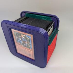May include: A purple 3D-printed deck box with a red textured side panel and a green interior accent. The front features a recessed frame holding a Magic: The Gathering card titled Curse of Marit Lage. The box contains a stack of black-edged cards.
