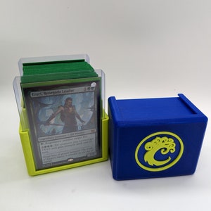 Peut inclure: Une boîte de deck bleue et jaune avec une carte Magic: The Gathering verte et blanche à l'intérieur. La carte est intitulée "Ezuri, Renegade Leader".