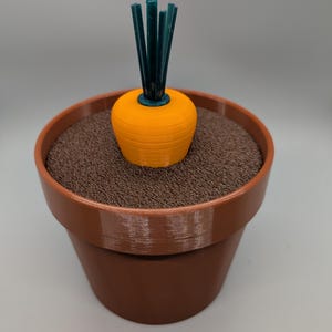以下が含まれることがあります： 緑色の茎が付いたオレンジ色の3Dプリントされたニンジンが、茶色の顆粒で満たされた茶色の植木鉢に入っています。 植木鉢も茶色で、明るい灰色の表面に置かれています。 ニンジンが焦点です。