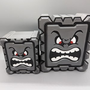 May include: Two gray 3D printed boxes with angry faces. The larger box has a wide open mouth with white teeth and red eyes. The smaller box has a smaller mouth with white teeth and red eyes.