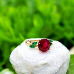 May include: A gold ring featuring a large, round, red ruby gemstone surrounded by smaller green gemstones. The ring has a delicate, leaf-like design.