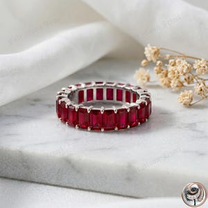 Op de afbeelding: Een zilveren ring met een doorlopende band van rechthoekige, dieprode edelstenen. De ring is geplaatst tegen een witte achtergrond met een takje gedroogde bloemen. De woorden "Full Moon Jewel" zijn zichtbaar op de achtergrond.