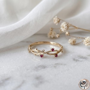 May include: A gold-colored ring with a delicate vine design. The ring features three small, round, red gemstones and one small, clear gemstone. The ring is set against a white background with a soft, blurred texture.