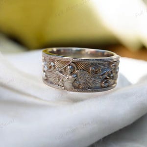 Puede incluir: Un anillo de plata con un diseño grabado que presenta un ángel sosteniendo una espada. El anillo tiene una banda ancha con tallas en relieve detalladas del ángel, alas y volutas decorativas. El anillo se muestra sobre una tela blanca.