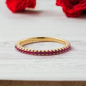 Op de afbeelding: Een gouden bandring met een doorlopende rij kleine, helderrode edelstenen. De ring ligt op een lichtgekleurd houten oppervlak, met een wazige rode bloem op de achtergrond. De ring is een delicaat sieraad.