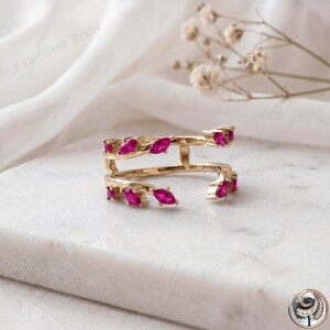 May include: A gold ring featuring a double band design, set with numerous marquise-cut, vivid pink gemstones. The ring is presented on a white surface. The text "Full Moon Jewel" is visible.