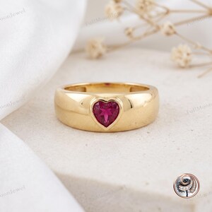 Pode incluir: Um anel de banda de ouro polido com uma única pedra preciosa rosa em forma de coração cravada no centro. O anel tem um perfil liso e arredondado com um acabamento brilhante, exibindo uma pedra rosa vibrante na banda de metal dourado.