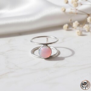 May include: A silver ring with a double-band design. The ring features a round, light pink opal gemstone set in the center. The ring is displayed on a white surface with soft lighting, showcasing the jewelry's delicate details.