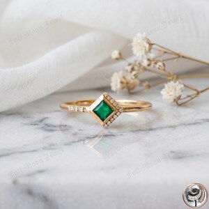 May include: A gold ring featuring a square emerald center stone, surrounded by a halo of small diamonds. Additional diamonds are set along the band. The ring is displayed on a marble surface with white fabric and dried flowers in the background.