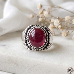 May include: A silver ring featuring a large, oval-shaped, deep red cabochon stone at its centre. The stone is surrounded by a twisted rope-style metal border and small silver spheres, set within a polished silver band with decorative side details.