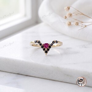 May include: A V-shaped gold ring featuring a central triangular pink gemstone surrounded by small round black stones. The band has a polished finish and follows a chevron silhouette, with the stones set along the top edge of the ring's point.