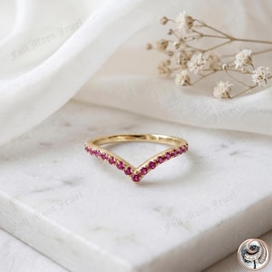 May include: A gold-colored V-shaped ring with a row of small, vibrant red gemstones. The ring is set against a white marble surface with a soft, white fabric in the background, and a sprig of white flowers.