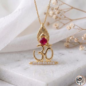 May include: Gold-toned pendant necklace featuring the Om symbol, adorned with a red gemstone and small white stones. The pendant hangs from a delicate chain. The necklace is displayed on a white surface.