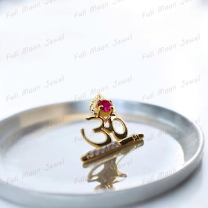 Könnte beinhalten: Ein goldfarbener Anhänger mit dem Om-Symbol, verziert mit einem roten Edelstein und kleinen weißen Steinen. Der Anhänger liegt auf einem silberfarbenen Teller. Der Text "Full Moon Jewel" wiederholt sich im Bild.