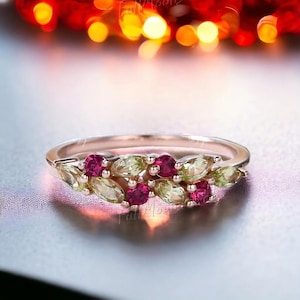 Può includere: Anello in oro rosa con un motivo floreale. Presenta pietre preziose rotonde, rosso intenso, e pietre ovali, verde chiaro, alternate. La fascia dell'anello è in metallo sottile e lucido.