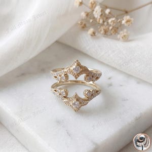 May include: Two gold-colored rings with intricate designs, featuring small clear gemstones. The rings have a crown-like shape with a central point and curved edges. The rings are displayed on a white surface with a soft, blurred background.