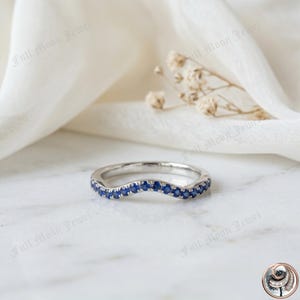 Pode incluir: Um anel curvo de prata com uma fileira de pequenas e vibrantes pedras preciosas azuis. O anel está sobre um fundo branco macio com sutis detalhes florais. O design do anel é elegante e moderno, adequado para várias ocasiões.