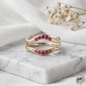 May include: A gold ring guard featuring two curved bands that meet in the center. Each band is set with a row of small, round, vibrant pink rubies. The metal has a polished finish, and the design creates a contoured shape to frame another ring.