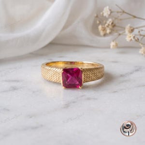 Può includere: Un anello in oro con una pietra preziosa rosa brillante dal taglio quadrato al centro. La fascia presenta una superficie testurizzata con un motivo a intreccio, che conferisce una finitura dettagliata al metallo attorno alla pietra rosa.