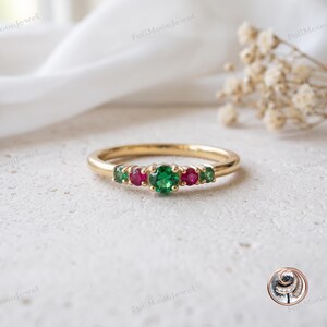 Puede incluir: Un anillo de oro con una piedra preciosa redonda verde central, flanqueada por dos piedras redondas rojas más pequeñas y dos piedras redondas verdes exteriores. El aro es liso y pulido, con una disposición de cinco piedras en engaste de garras.