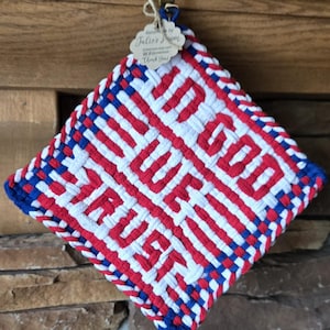 May include: A woven hot pad in red, white, and blue, featuring the phrase "In God We Trust." The square pad has a blue border and is made of woven fabric. A small tag is attached to the top.