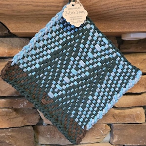 May include: Handmade woven pot holder in a square shape. The pot holder features a woven pattern in shades of blue, green, and brown. A tag attached to the top reads "Handmade by Julie's Loom" and includes a website and social media handles.