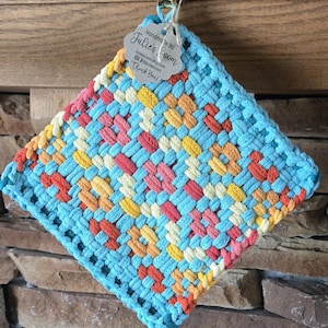 May include: A square woven potholder featuring a multi-colored pattern of light blue, yellow, orange, and pink yarn. A scalloped paper tag is attached to the top, printed with the text: Handmade by Julie's Loom, JuliesLoom.etsy.com, @juliesloom, Thank You!