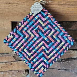 May include: A square woven potholder featuring a geometric maze pattern in navy blue, light blue, coral pink, and pale yellow. A white scalloped tag is attached to the top, printed with the text Handmade by Julie's Loom and social media icons.